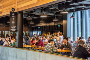 A cafe inside a building. The interior is made up of wood and black along with concrete. A large number of visitors are seen sitting at tables in the cafe.