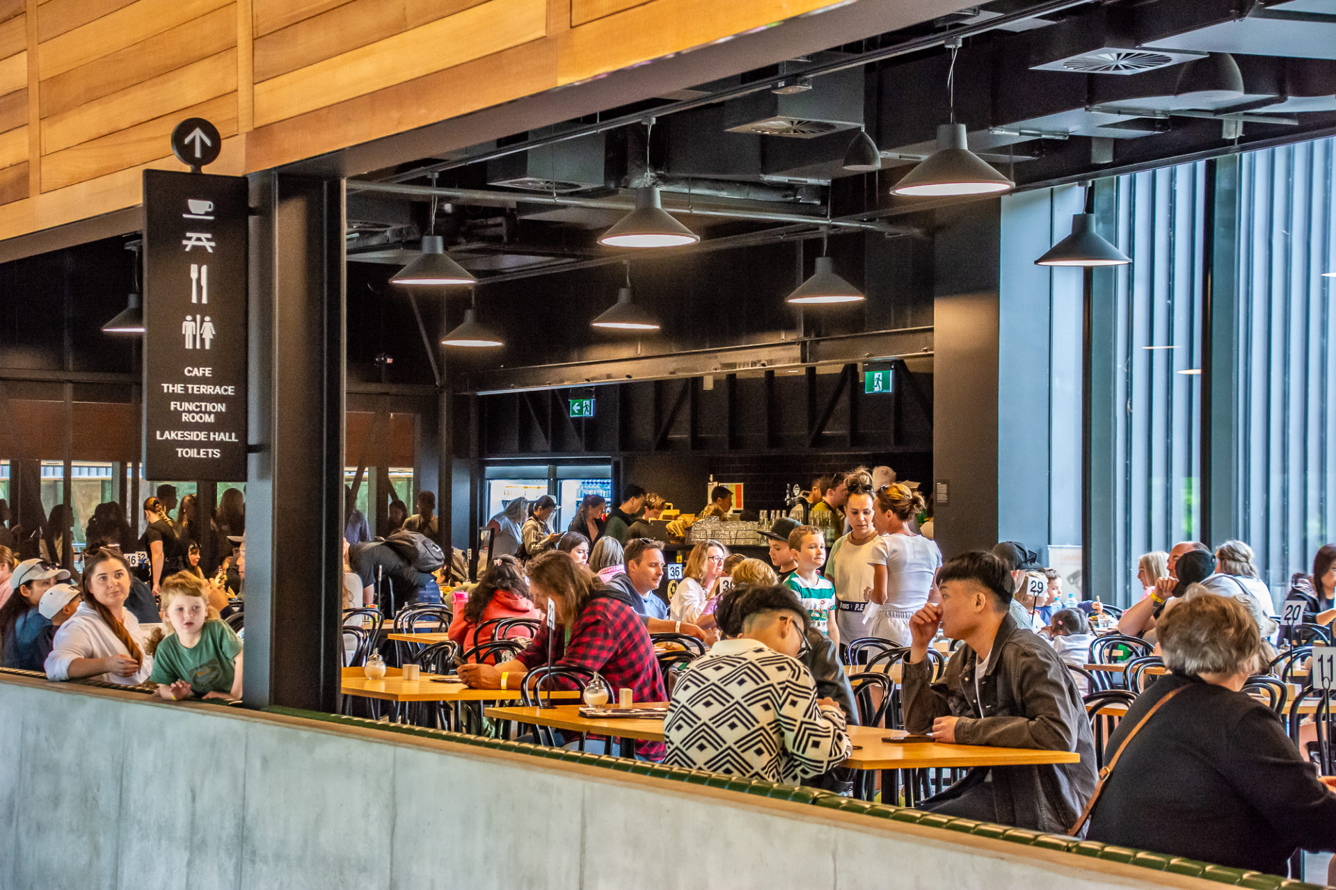A cafe inside a building. The interior is made up of wood and black along with concrete. A large number of visitors are seen sitting at tables in the cafe.
