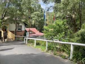 Paved pathway to station from Accessible Car Parks. The pathway is bitumen but steep. It has white, wooden handrails. The path turns right on it's way to the Belgrave booking office. A sign directs passengers to follow the path to the right to check in.