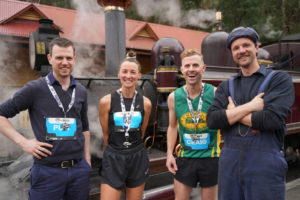 Four adults stand in front of a locomotive. Three of them have running clothes on, while the fourth is dressed in dark blue overalls.