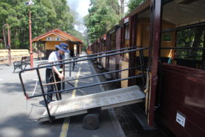 A ramp is attached to one of Puffing Billy's accessible carriages with open doors. A Puffing Billy volunteer can be seen operating the ramp to put it in place.