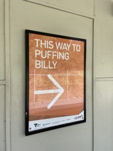 Sign at the Belgrave Metro Train station showing the way to Puffing Billy Railway. The sign has an orange background with white text saying "This way to Puffing Billy" with an arrow.