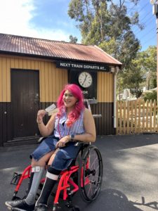 A woman in a wheelchair in front of a yellow and brown painted wooden building. An old-style clock can be seen in the background, showing the departure of the next train. The woman is holding her Puffing Billy tickets and smiling at the camera.