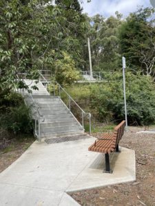 Concrete Steps From Top Lakeside Car Park. The steps has handrails and there is a wooden bench at the bottom of the steps. A concrete path continues on.