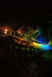 Puffing Billy Train of Lights traverses the trestle bridge over Cockatoo Creek. The locomotive and carriages are illuminated in rainbow colours.
