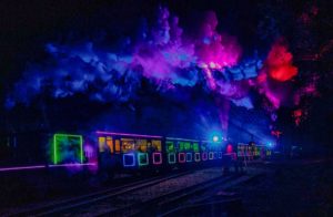 A illuminated steam train in the darkness. The train and steam are lit up in purple, pink, red, blue and green hues.