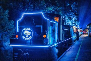 An illuminated train with blue hues sits at a platform. The front of the locomotive has a headboard saying Puffing Billy Railway Train of Lights. The blue hues illuminate the trees in the background.