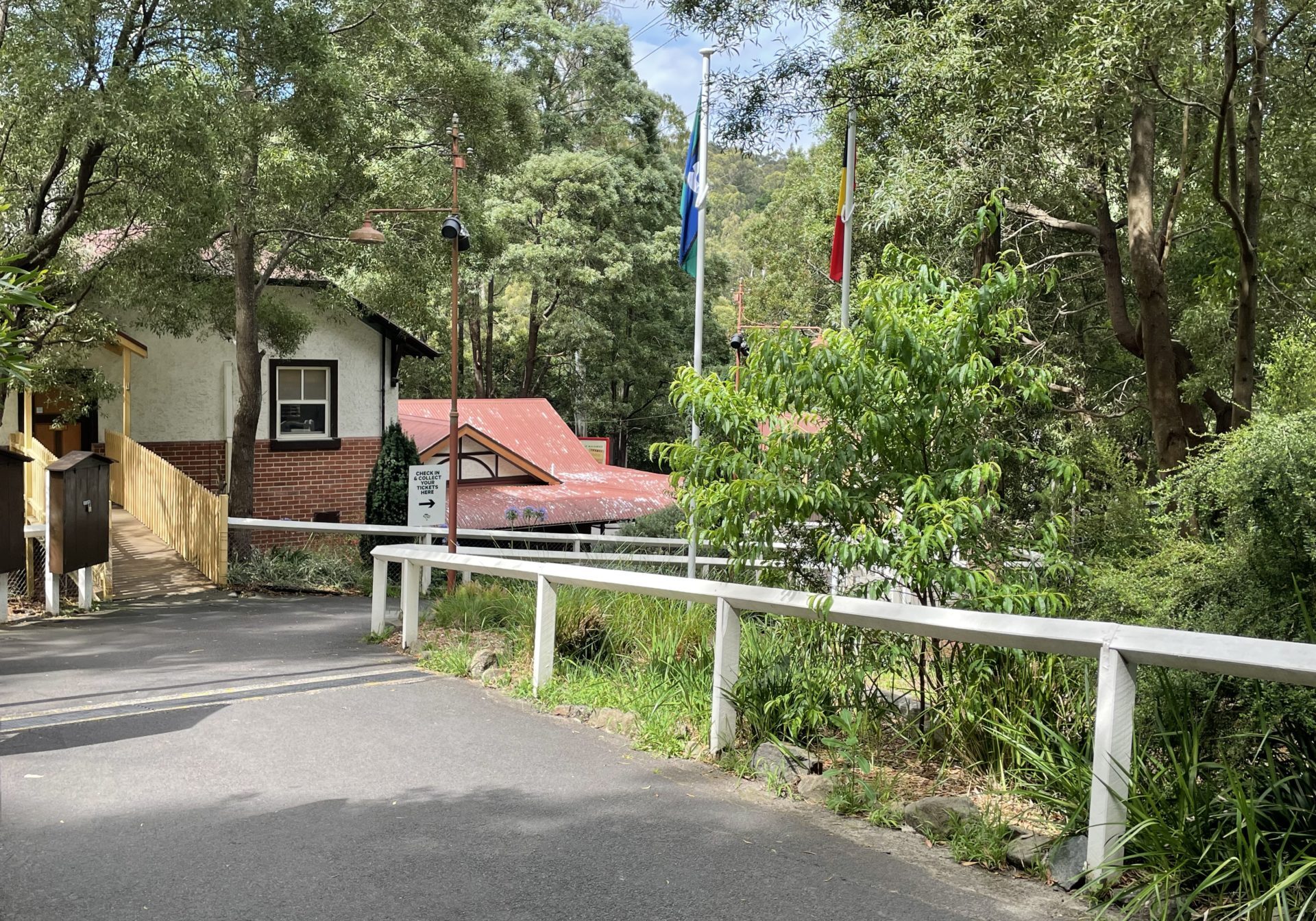 Paved pathway to station from Accessible Car Parks. The pathway is bitumen but steep. It has white, wooden handrails. The path turns right on it's way to the Belgrave booking office. A sign directs passengers to follow the path to the right to check in.