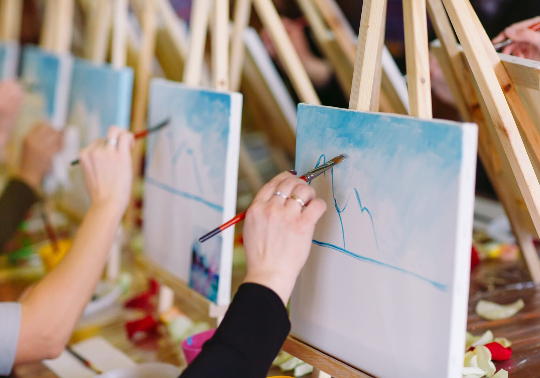 Visitors take part in a Paint and Sip Workshop. They are using wooden easels placed side by side at a table and are painting on canvases.