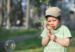 A boy is blowing bubbles with a bubble wand made out of red pipe cleaner. The pipe cleaner is made into a heart shape. The boy is wearing a light green t-shirt and beige cap and is looking intently at the wand. Greenery can be seen in the background.